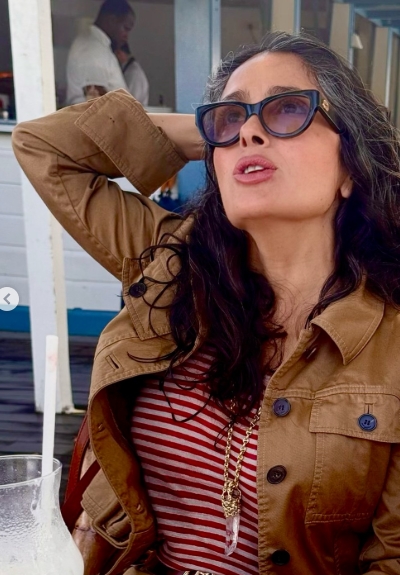 Salma Hayek in a red and white striped tee and tan utility jacket, accessorized with sunglasses and a crystal pendant.