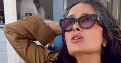 Salma Hayek wearing a printed bikini top and white linen pants on a yacht at sunset.