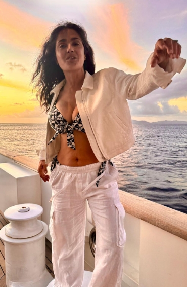 Salma Hayek wearing a printed bikini top and white linen pants on a yacht at sunset.
