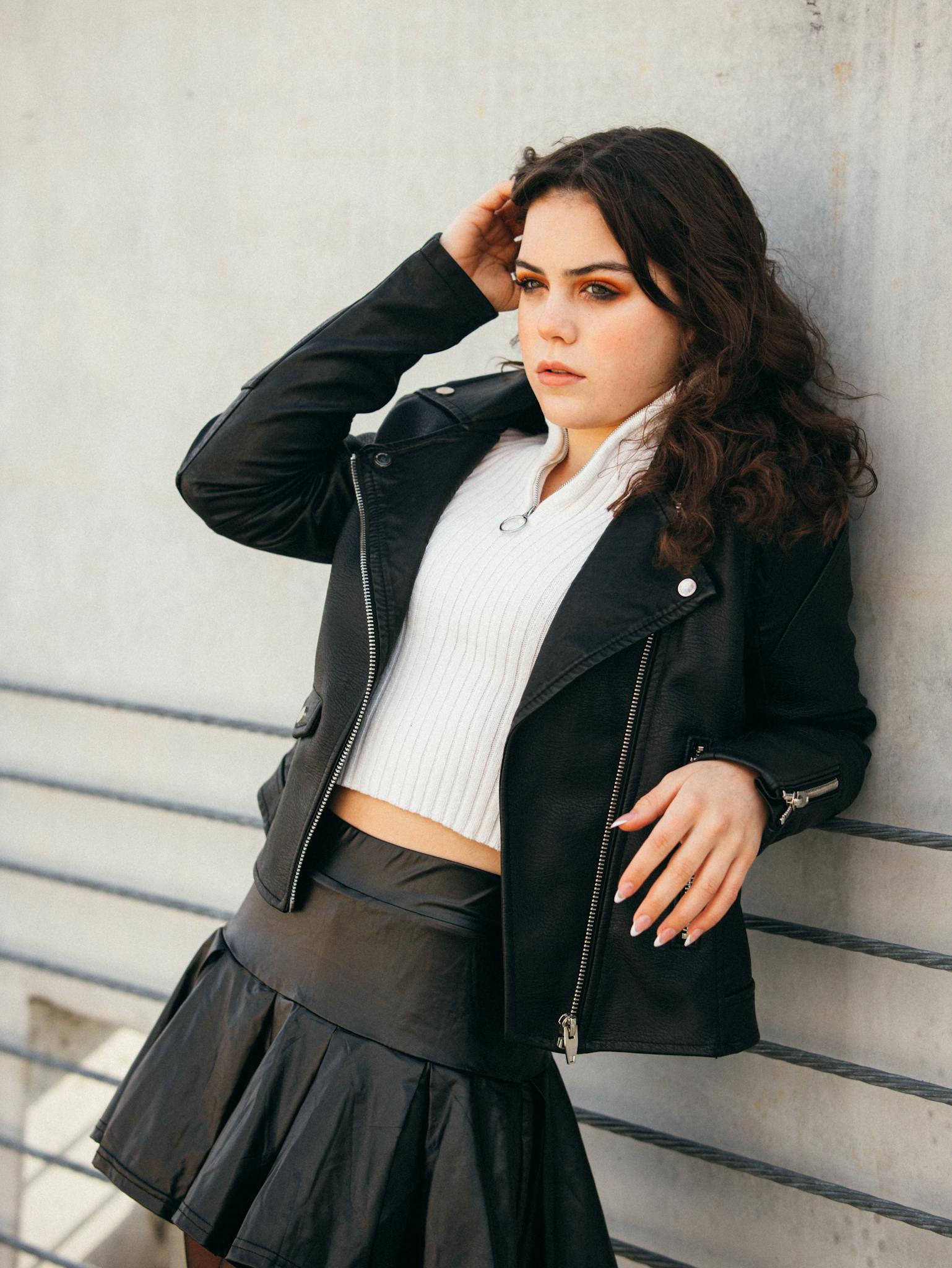 Fashionable woman in a black leather jacket leaning against a wall outdoors.